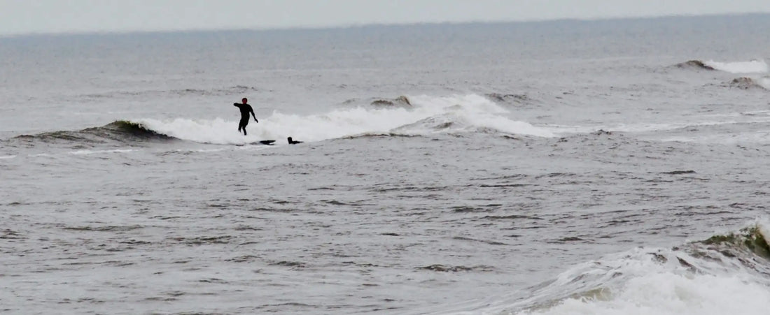 Imagem de mar aberto com a silhueta de um surfista no mar ao fundo. A imagem é de um final de tarde e está em preto e branco, com algumas ondas baixas no mar.