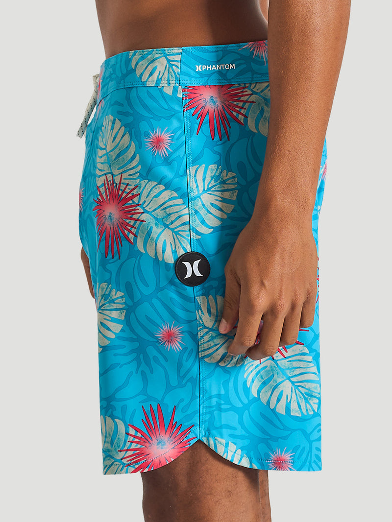 Boardshort azul com estampa tropical de folhas bege e flores vermelhas, cós ajustável por cordão e logo da marca na lateral esquerda.