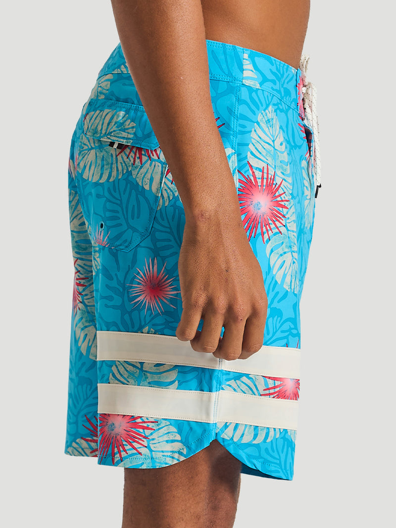 Boardshort azul com estampa tropical de folhas e flores vermelhas, detalhe de listras brancas na parte inferior e bolso traseiro com aba.