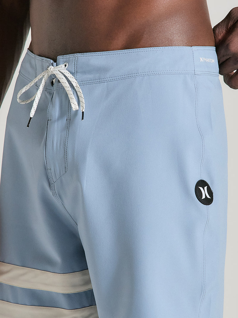 Close de boardshorts phantom 18 polegadas azul com cordão branco ajustável, logo circular preto na lateral e listras horizontais no lado direito.