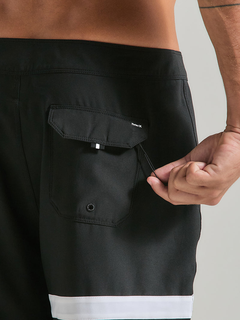 Close de boardshorts phantom 18 polegadas preto, com bolso traseiro fechado por uma aba e cordão. Detalhe de faixa branca na barra.