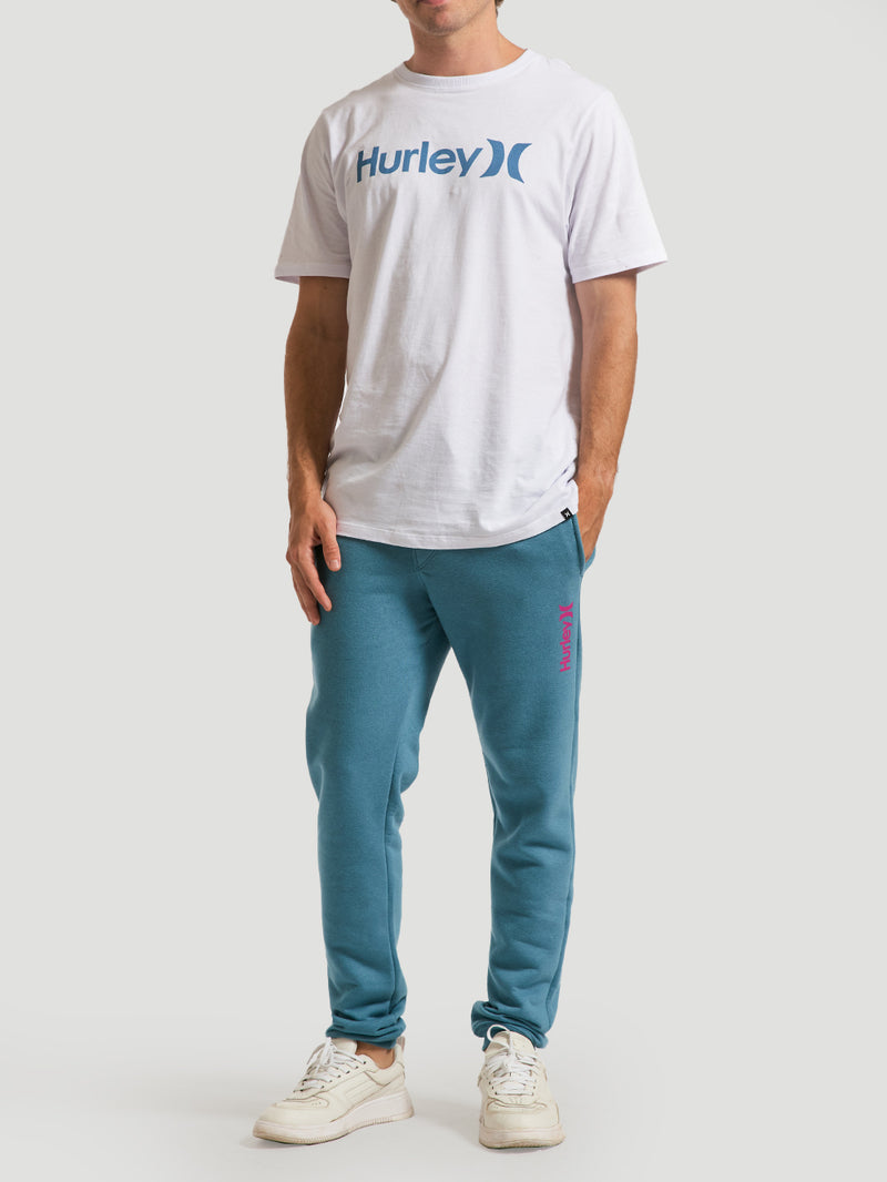 Homem veste uma calça moletom azul básica com modelagem ajustada, logo rosa na perna, combinada com camiseta branca estampada e tênis brancos.