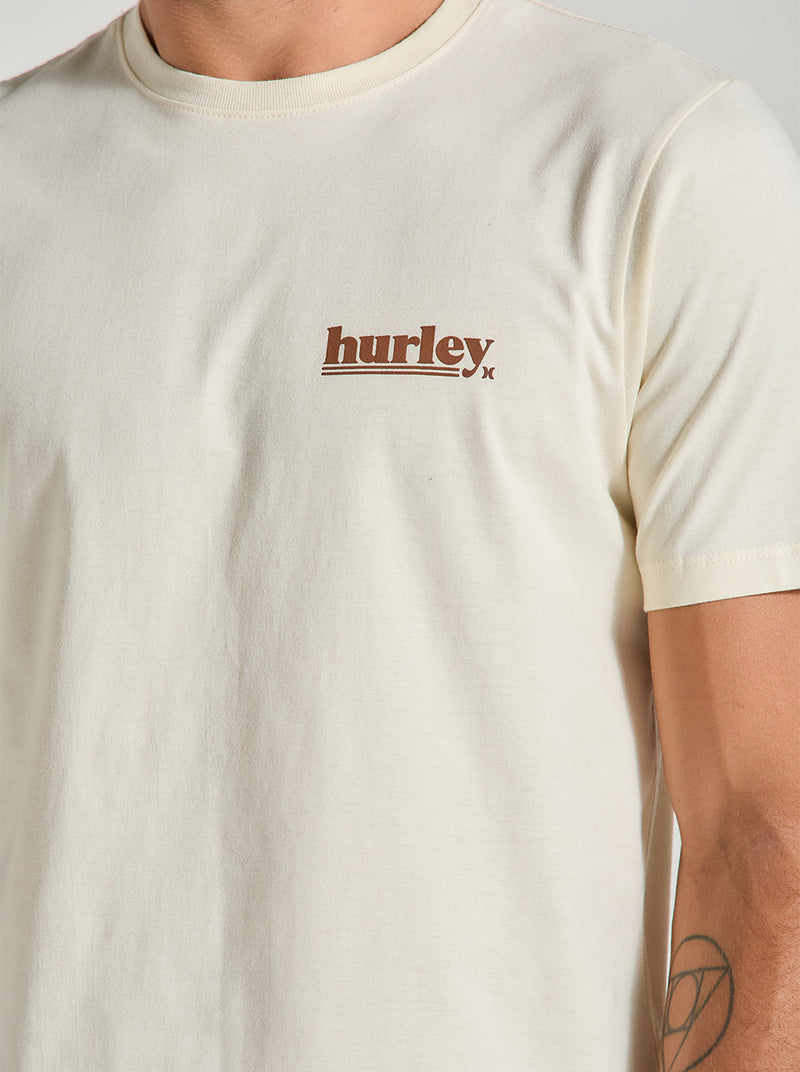 Close da camiseta algodão básica off white, com gola redonda e mangas curtas. Possui o logotipo "hurley" em marrom no lado esquerdo do peito.