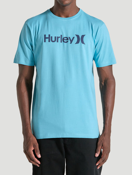 Homem veste uma camiseta azul manga curta com estampa centralizada Hurley em letras escuras, combinada com calça preta, em fundo cinza claro.