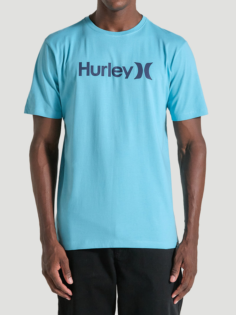 Homem veste uma camiseta azul manga curta com estampa centralizada Hurley em letras escuras, combinada com calça preta, em fundo cinza claro.