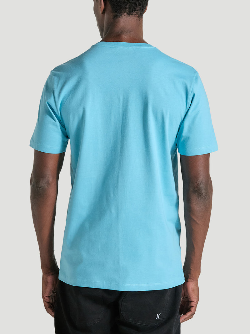 Homem de costas veste uma camiseta azul manga curta com tecido liso, caimento reto e combinação com calça preta, em fundo cinza claro.