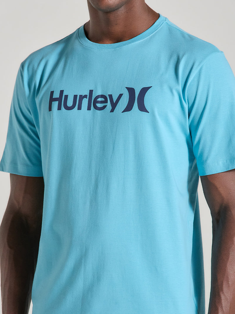 Homem veste uma camiseta azul manga curta com estampa frontal azul-marinho Hurley, tecido liso e caimento reto, em fundo neutro.