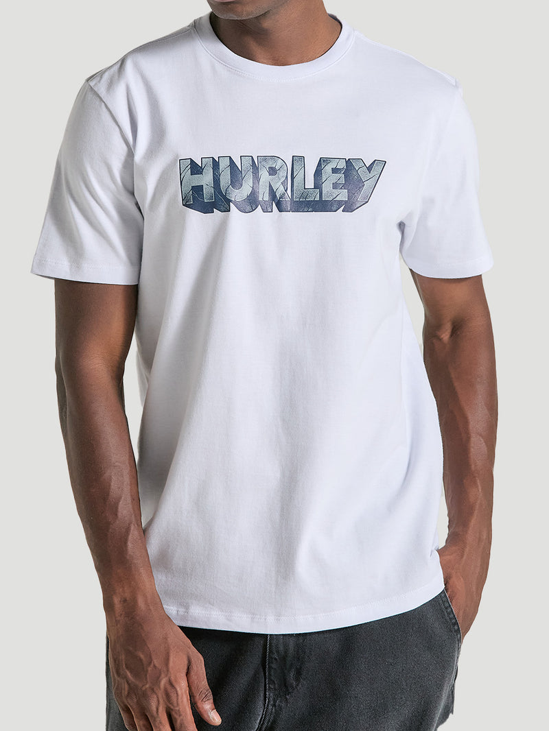 Homem vestindo camiseta branca manga curta estampada com gola redonda. O destaque é o logotipo Hurley em grande, com textura azul e efeito 3D.