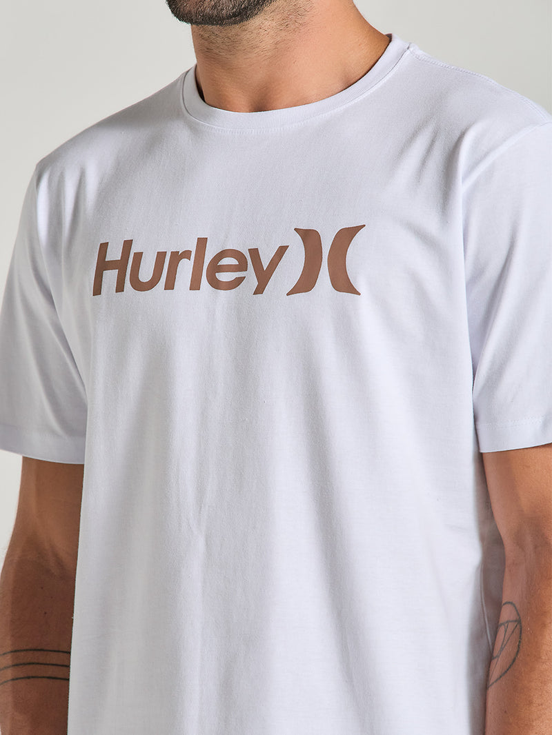 Homem veste uma camiseta branca manga curta com estampa Hurley discreta em marrom, tecido liso e caimento reto, em fundo claro e neutro.