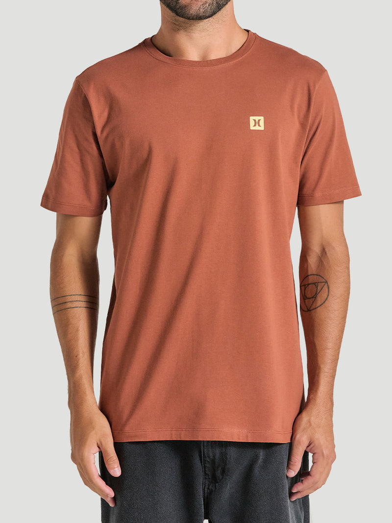 Camiseta em algodão marrom com gola redonda e mangas curtas. A camiseta possui um pequeno logo amarelo no lado esquerdo do peito.