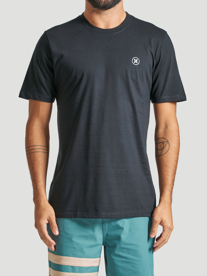 Homem veste camiseta em algodão preta com gola redonda, caimento reto e pequena logo branco no lado esquerdo do peito, combinada com bermuda.