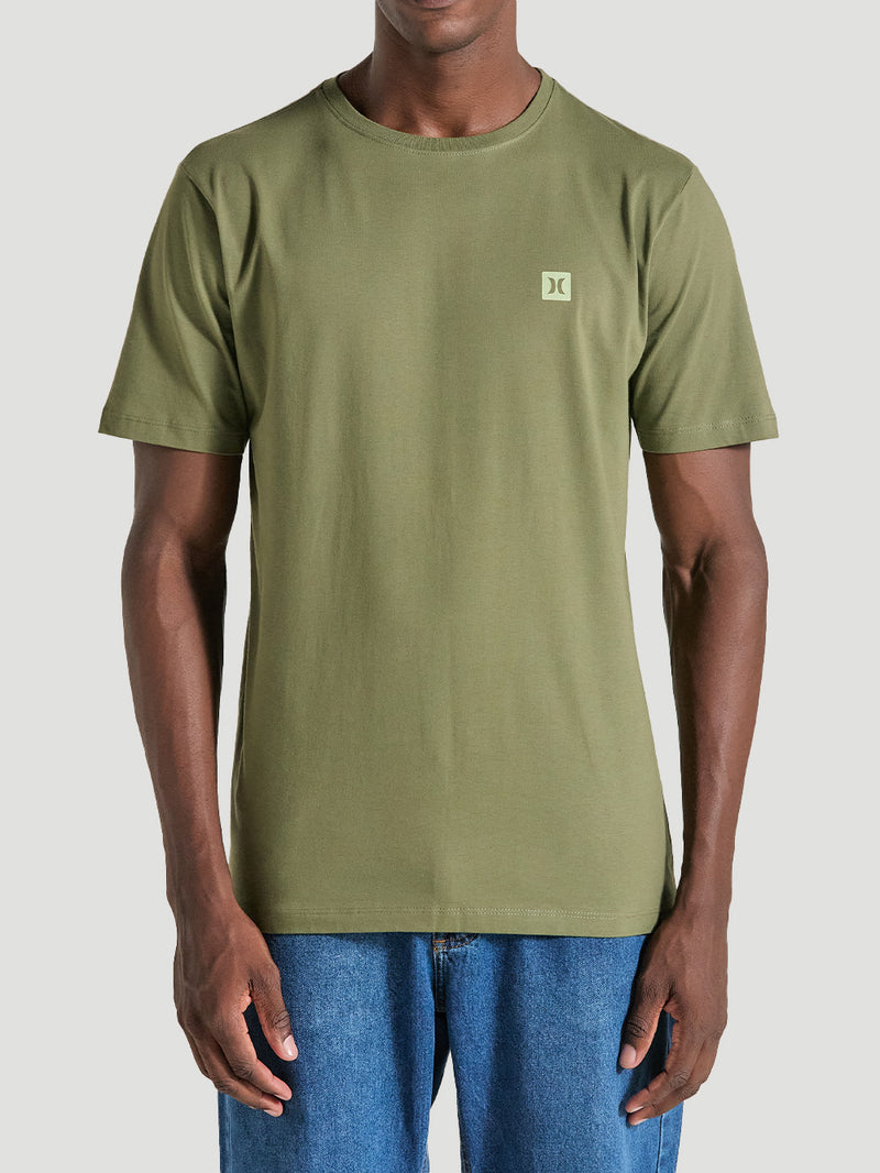 Homem vestindo camiseta em algodão verde militar de gola redonda e mangas curtas, com logotipo pequeno no peito esquerdo, combinada com jeans.