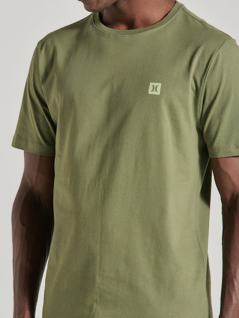 Homem usando uma camiseta em algodão verde militar com gola redonda e mangas curtas. A camiseta apresenta um logotipo discreto no lado esquerdo do peito.