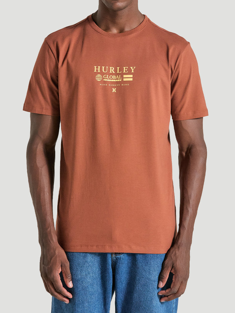 Homem veste camiseta marrom estampa frontal manga curta com escrita "Hurley Global Trademark" em amarelo, combinada com calça jeans azul casual.