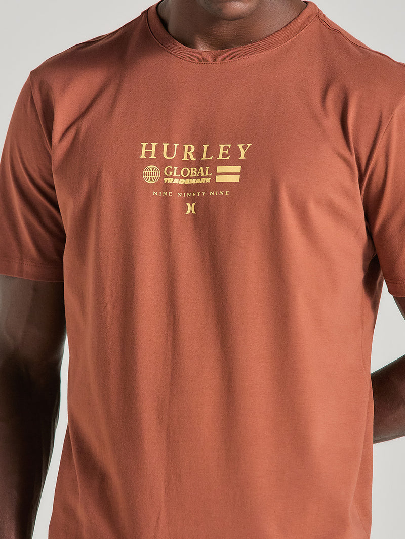 Homem veste camiseta marrom estampa frontal manga curta e dizeres em amarelo. O design apresenta o texto "Hurley Global Trademark" e elementos gráficos minimalistas.
