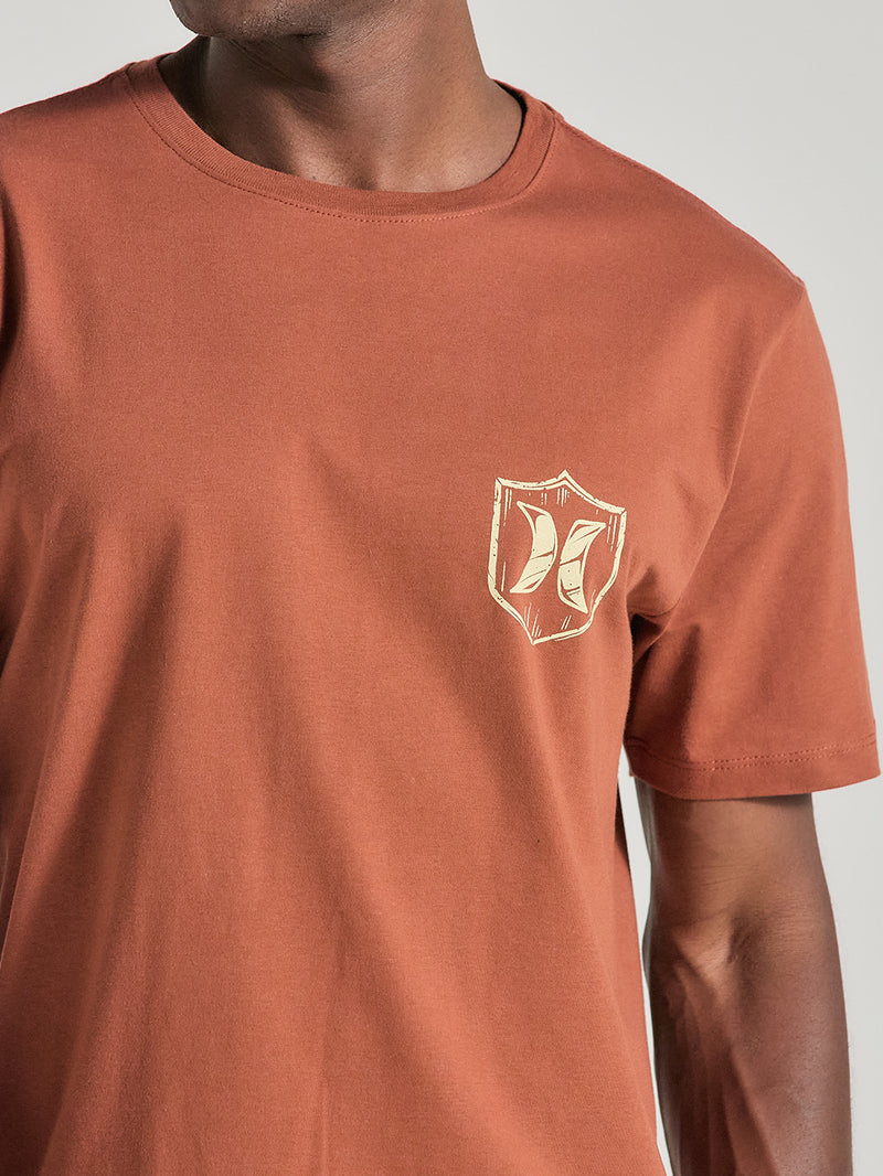 Camiseta marrom manga curta estampa pequena no lado esquerdo do peito, apresentando um escudo bege estilizado com o logotipo central.