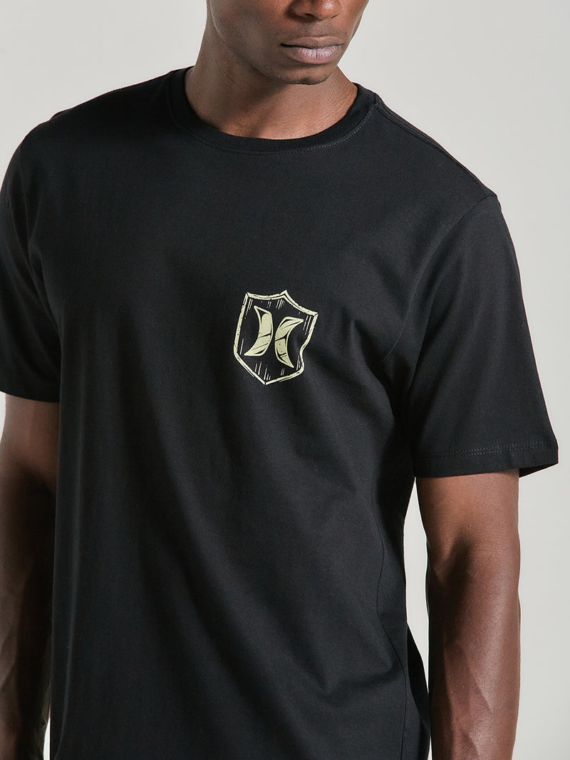 Homem usando camiseta preta manga curta estampa com arte pequena no lado esquerdo do peito, em formato de escudo com o logotipo "Hurley".