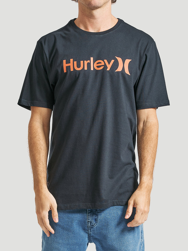 Homem veste uma camiseta preta manga curta com estampa Hurley frontal em laranja, combinada com jeans azul claro, em fundo neutro.