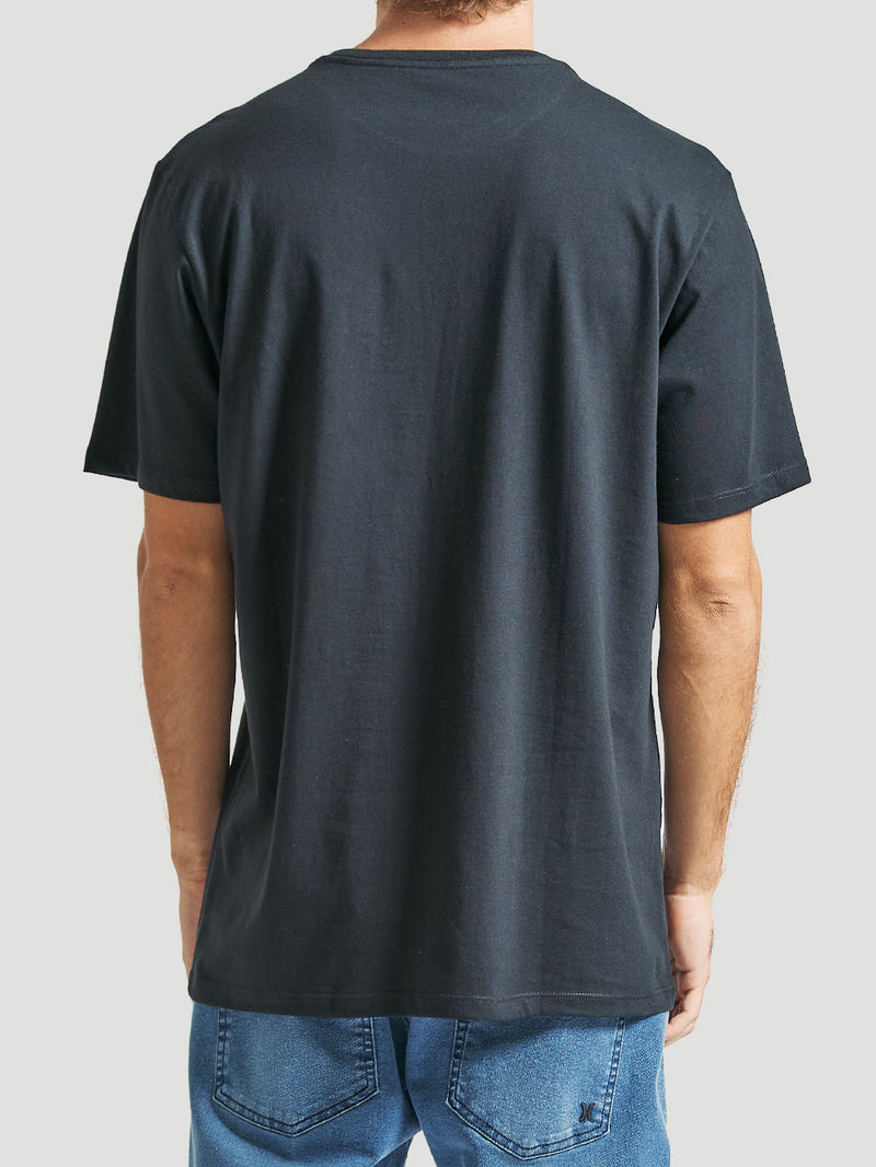 Homem de costas veste uma camiseta preta manga curta com caimento reto e tecido liso, combinada com jeans azul claro, em fundo cinza claro.