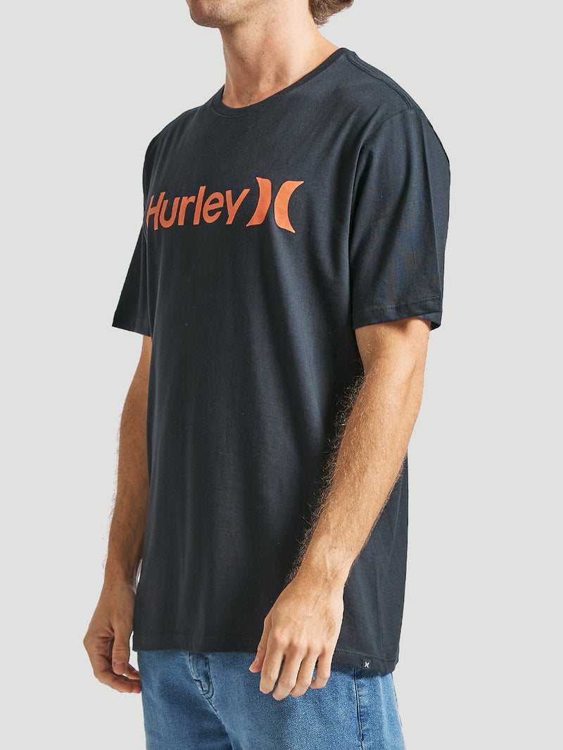 Homem veste uma camiseta preta manga curta com estampa frontal laranja Hurley, tecido liso e caimento reto, combinada com jeans azul, em fundo claro.