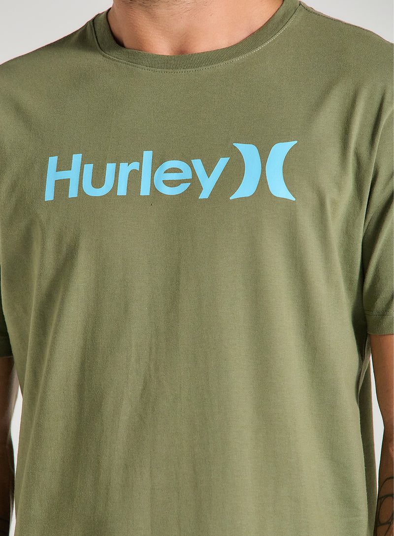 Homem veste uma camiseta verde manga curta com estampa frontal azul claro Hurley, de tecido liso e caimento reto, em fundo claro.