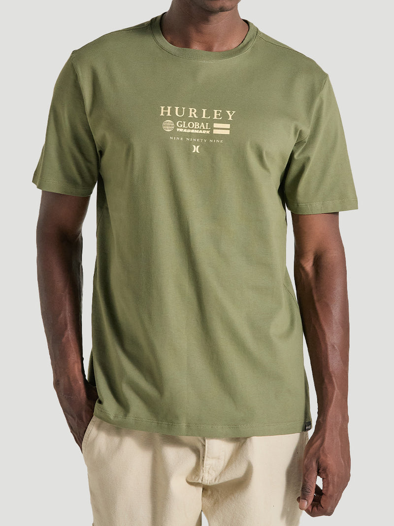 Homem vestindo uma camiseta verde militar estampa frontal manga curta com escrita em texto bege e calça bege, em fundo neutro.