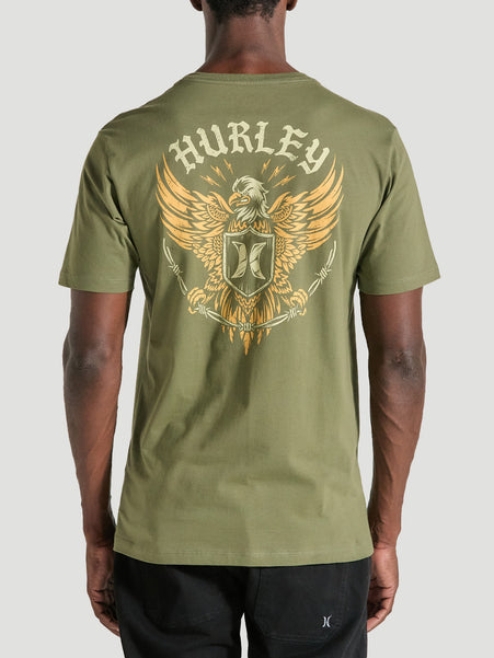 Homem vestindo camiseta verde militar manga curta estampa nas costas, apresentando uma águia dourada com asas abertas, escudo central e o texto "Hurley".