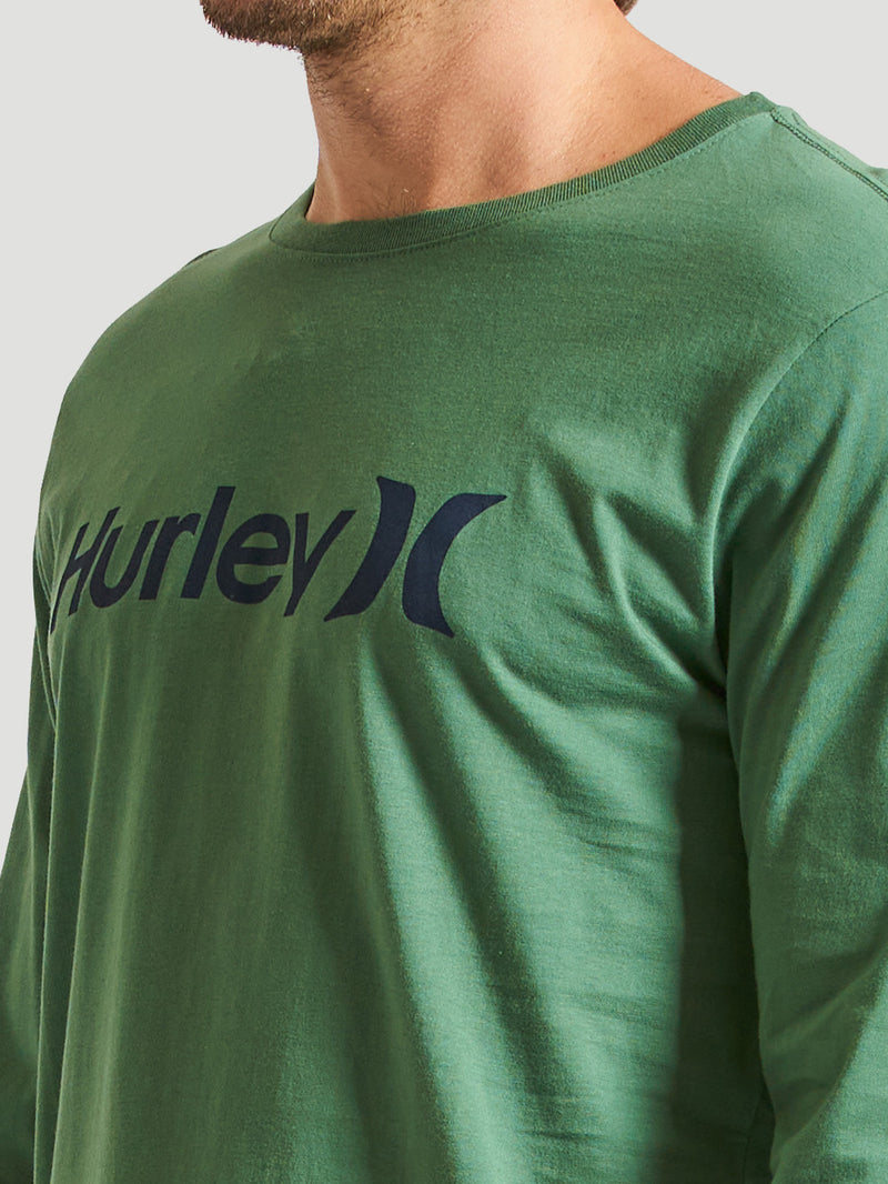 Homem vestindo camiseta verde militar manga longa com estampa frontal azul escura do logo "Hurley". Enquadramento lateral mostrando ombro e peito. Fundo claro.