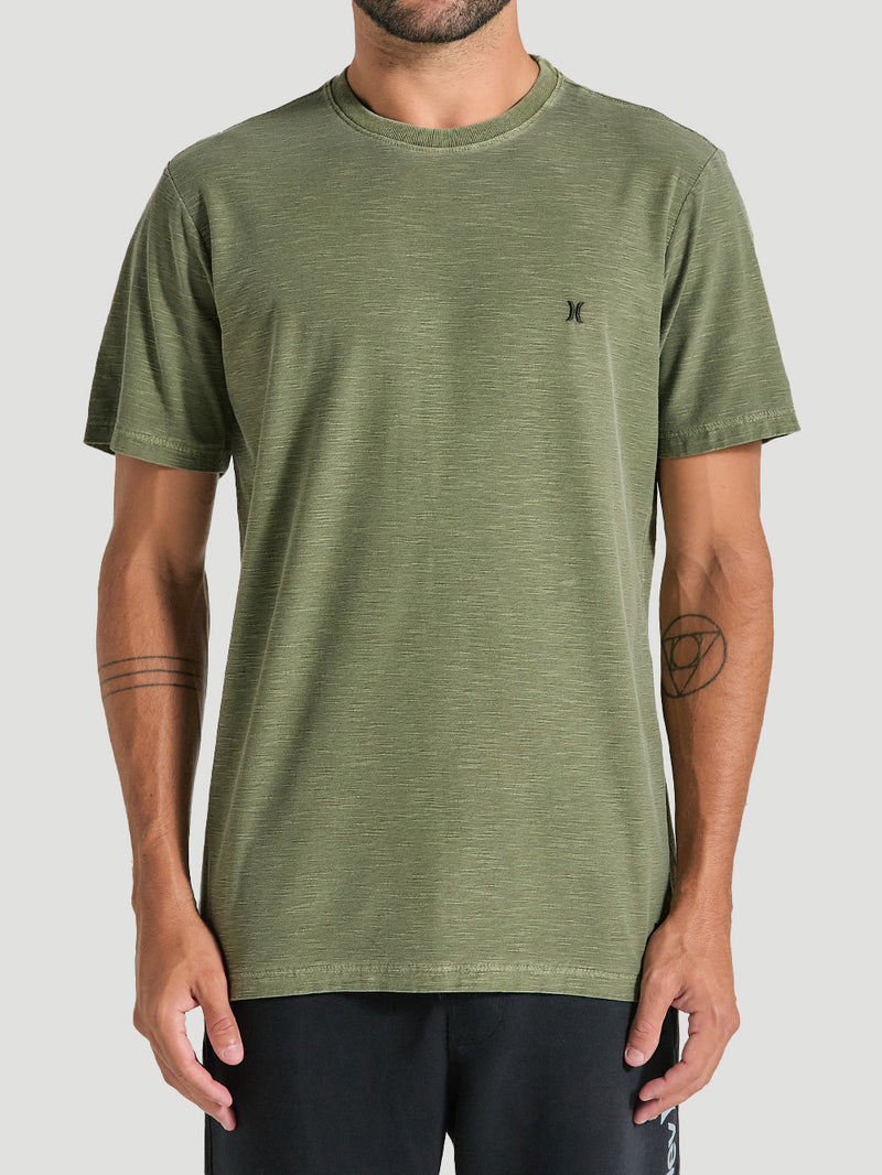 Homem usando a camiseta verde militar tinturada manga curta com tecido texturizado e pequeno logo preto no lado esquerdo do peito. Fundo cinza claro.