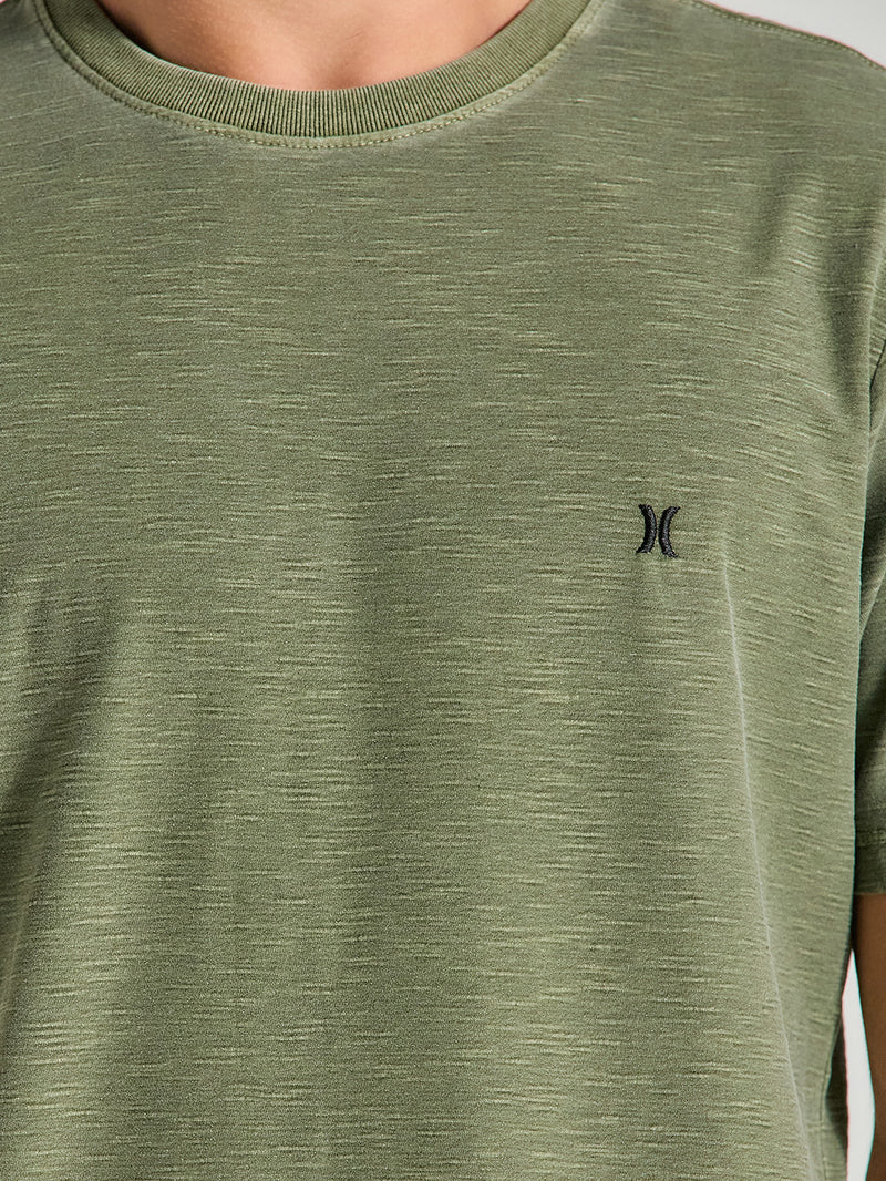 Close da camiseta verde militar tinturada manga curta com textura leve no tecido e pequeno logo preto bordado no lado esquerdo do peito.