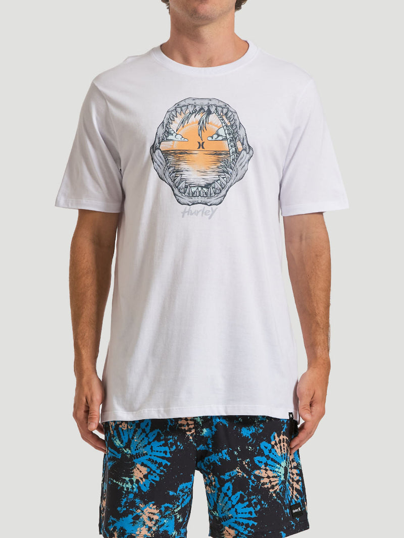 Homem usa camiseta estampada algodão branca com arte central destacada de um pôr do sol visto através de mandíbulas abertas, criando um design tropical e ousado.