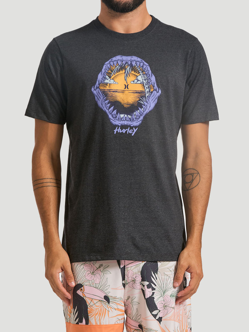 Homem usa camiseta estampada algodão preta com ilustração central de mandíbulas abertas revelando um pôr do sol tropical. Combina com bermuda floral colorida.