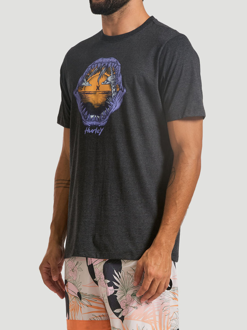 Homem veste camiseta estampada algodão preta com arte colorida central de mandíbulas abertas revelando um pôr do sol tropical, combinada com bermuda floral vibrante.