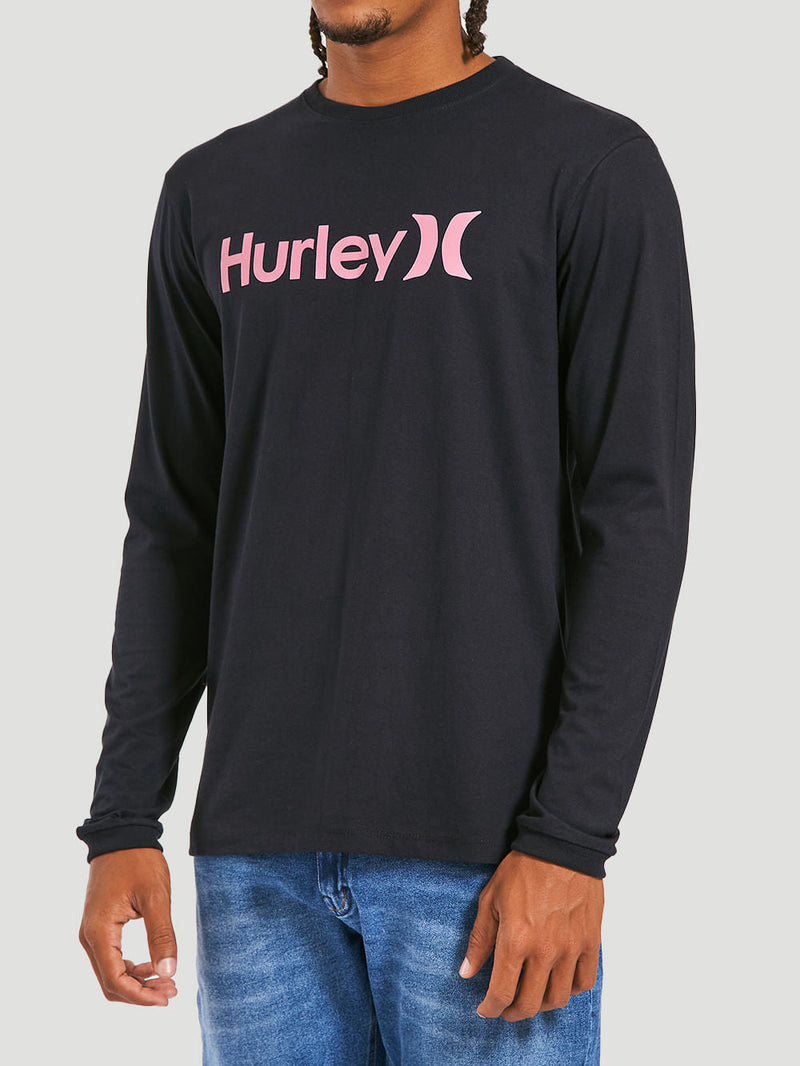 Homem usando a camiseta preta manga longa com estampa frontal rosa escrita "Hurley", combinada com calça jeans azul claro. Fundo neutro.