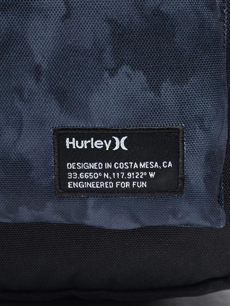 Detalhe da mochila Hurley com etiqueta preta contendo o logotipo branco, coordenadas geográficas e a frase "Engineered for fun" impressa.