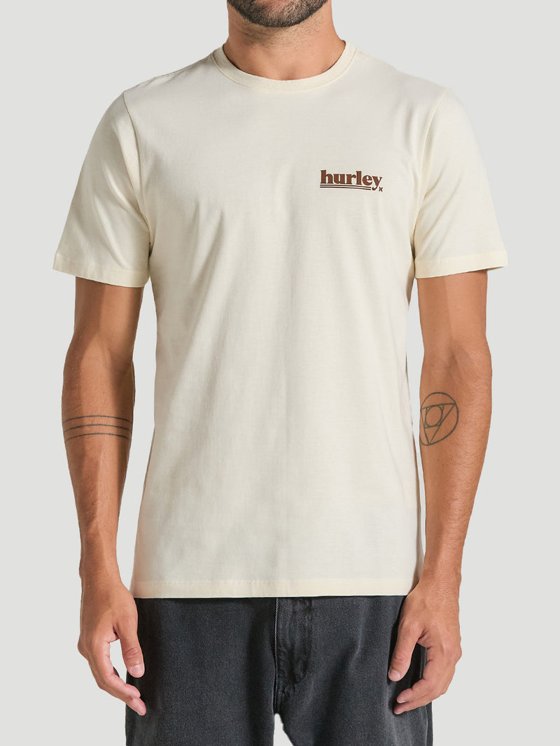 Homem vestindo uma camiseta algodão básica off white com gola redonda e mangas curtas. Logotipo "hurley" marrom destacado no lado esquerdo do peito.