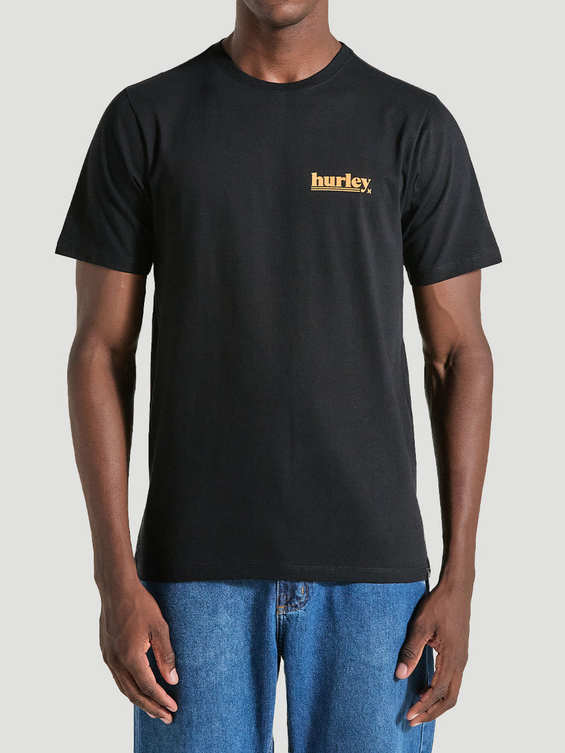Homem vestindo uma camiseta algodão básica preta com gola redonda e mangas curtas. O design simples inclui um pequeno logotipo amarelo no lado esquerdo do peito.
