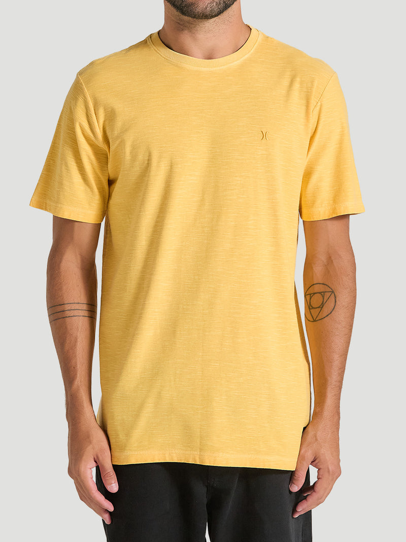 Camiseta amarela tinturada manga curta com pequeno logo bordado no peito, combinada com calça preta. Fundo neutro.