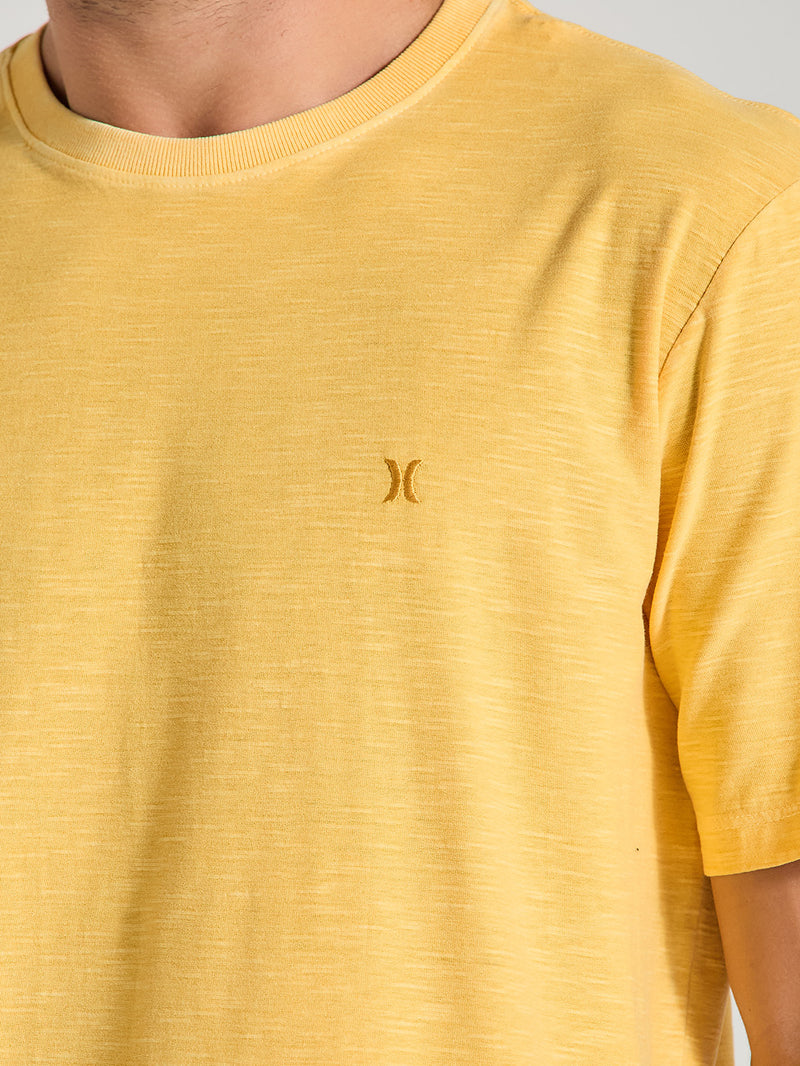 Detalhe da camiseta amarela tinturada  manga curta com logo discreto bordado em amarelo no peito. Fundo neutro claro.