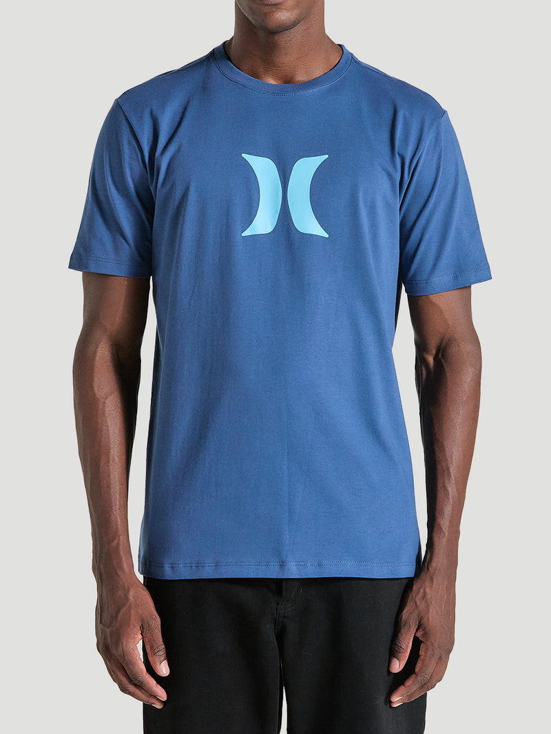 Homem veste uma camiseta azul manga curta básica com estampa minimalista azul clara no centro, tecido liso e caimento reto, combinada com calça preta.