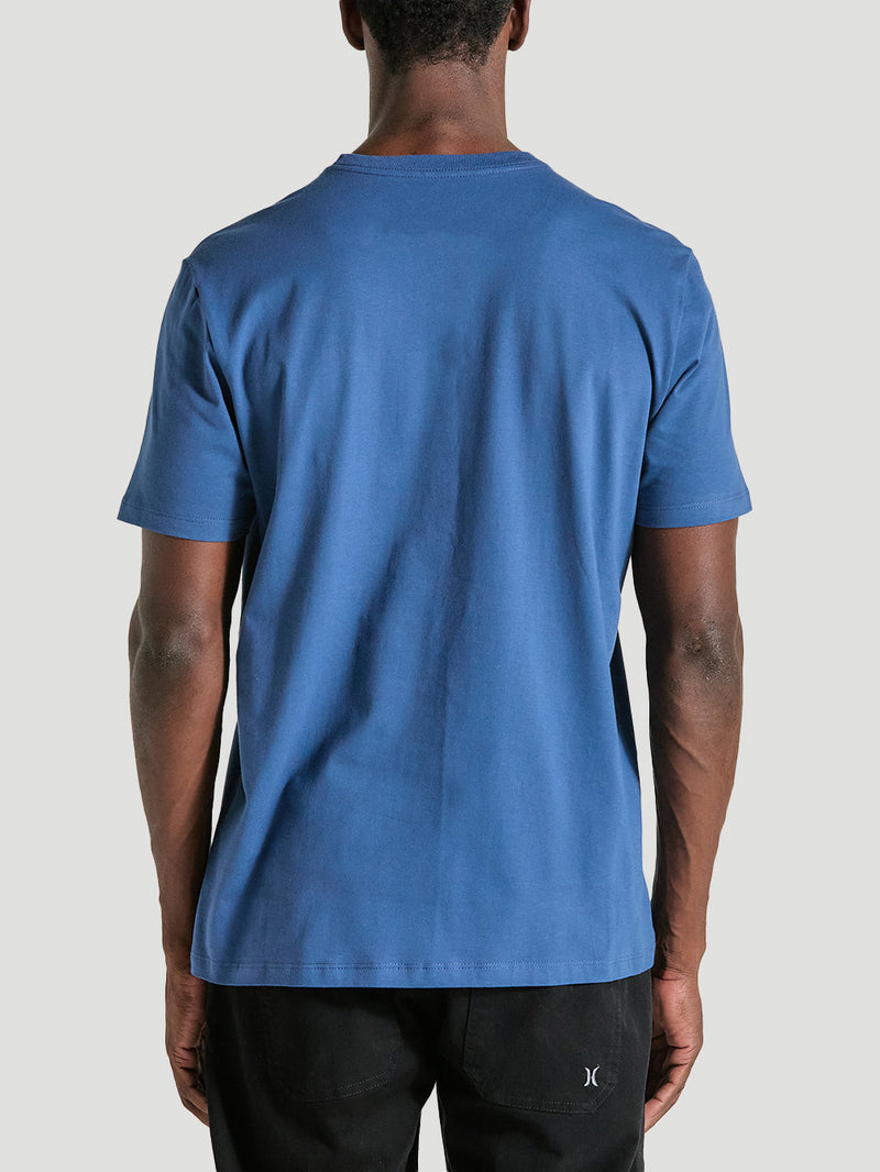 Homem de costas veste uma camiseta azul manga curta básica de tecido liso, caimento reto e gola redonda, combinada com calça preta, em fundo cinza claro.