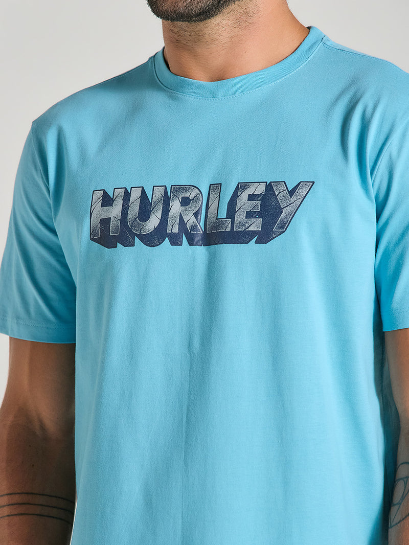 Homem vestindo camiseta azul manga curta estampada. A estampa frontal é do logotipo "Hurley" em letras estilizadas escuras.