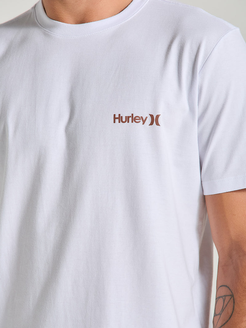 Homem vestindo uma camiseta branca com estampa, gola redonda e mangas curtas. O detalhe é o logotipo Hurley em marrom no lado esquerdo do peito.