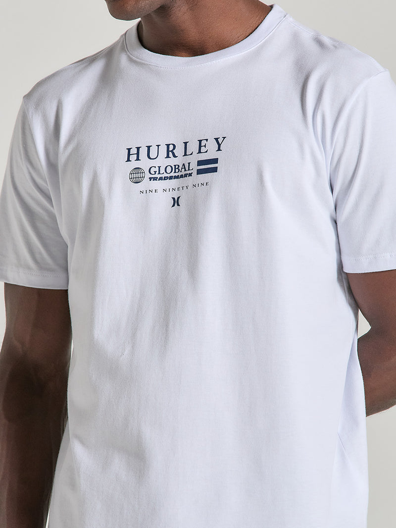 Homem veste camiseta branca estampa frontal manga curta de algodão com escrita em azul escuro. O design inclui o texto "Hurley Global Trademark". Estilo casual.