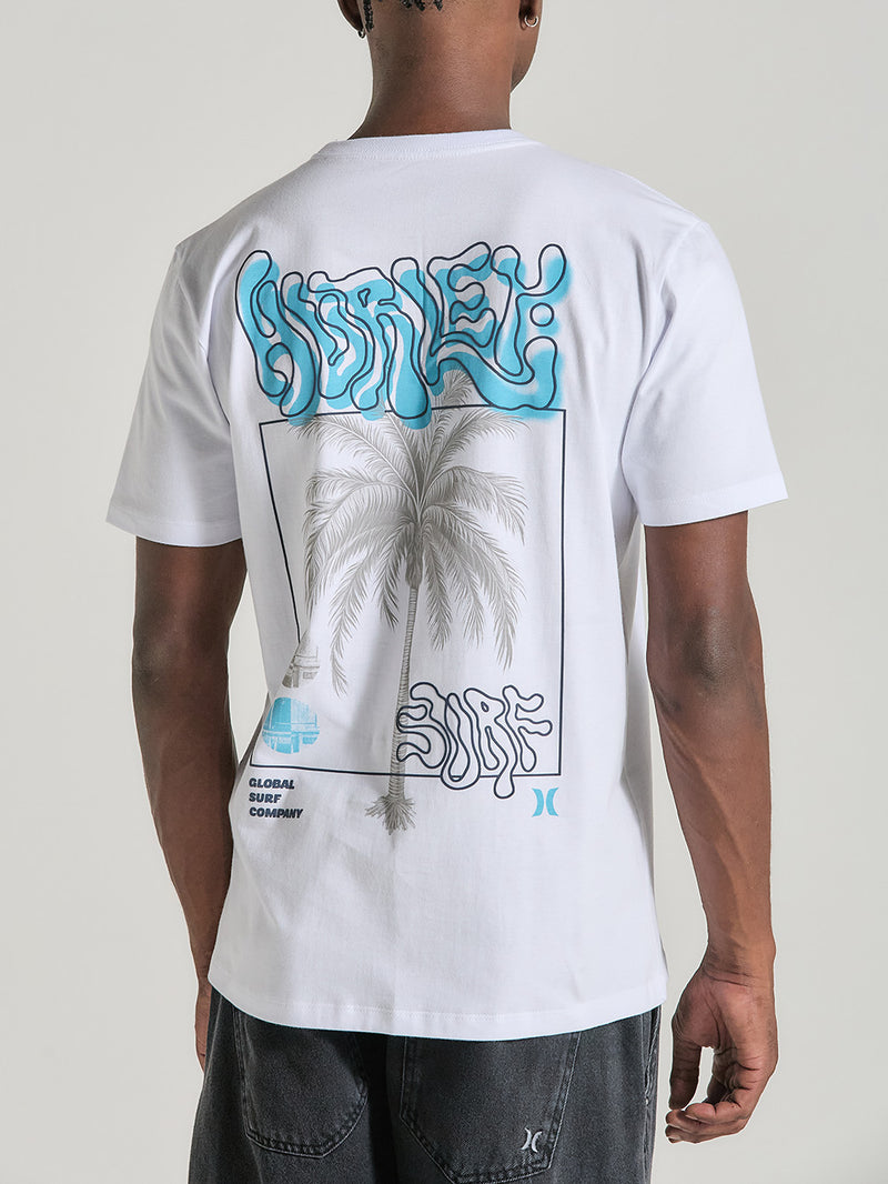 A camiseta branca manga curta estampa relevo na imagem apresenta uma estampa estilizada nas costas, envolvendo o tema surf. A arte inclui palmeiras, texto desenhado com uma fonte fluida e um elemento gráfico central com a palavra "Surf" em destaque.