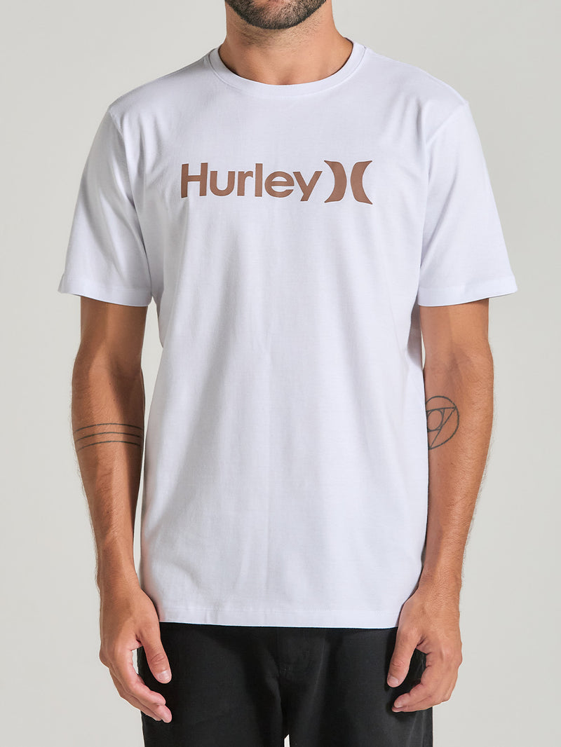 Homem veste uma camiseta branca manga curta com estampa centralizada, combinada com calça preta. Fundo neutro e iluminação suave.