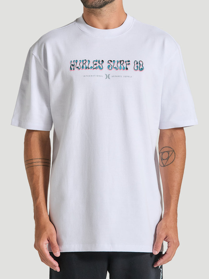 Homem vestindo camiseta de algodão oversized branca manga curta com estampa “Hurley Surf Co.” no centro, combinada com calça preta.