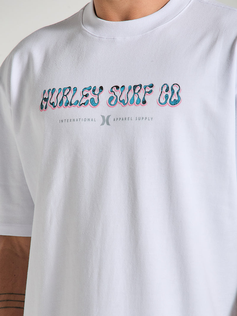 Close-up de uma camiseta de algodão oversized branca manga curta, com estampa central "Hurley Surf Co." em estilo colorido e letras menores abaixo.