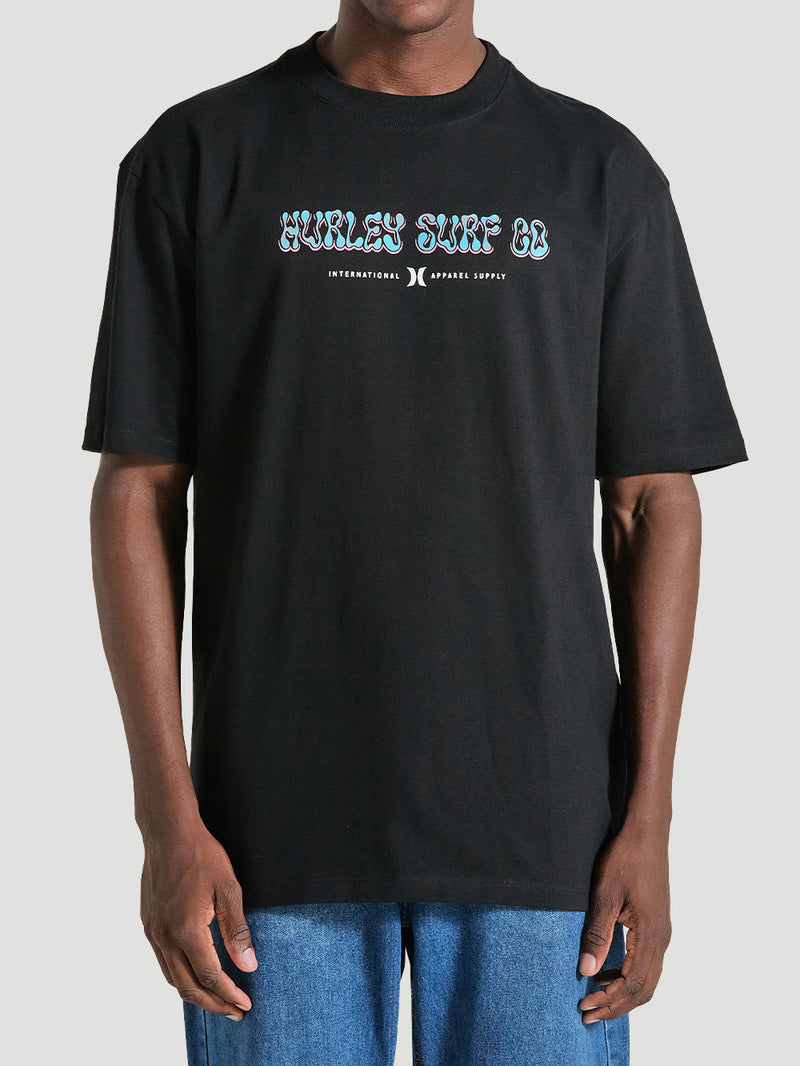 Homem vestindo camiseta de algodão oversized preta manga curta com estampa "Hurley Surf Co." colorida no peito, combinada com calça jeans azul.