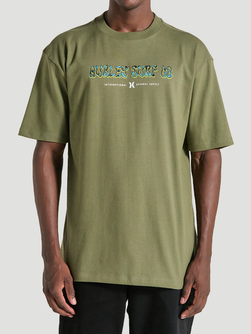 Homem vestindo camiseta de algodão oversized verde militar manga curta com estampa "Hurley Surf Co." no peito, combinada com bermuda preta casual.